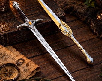 Puede incluir: Una espada plateada con mango negro y una piedra preciosa color ámbar engastada en la guarda. A su lado yace una vaina ornamentada a juego con intrincados detalles de filigrana en oro y plata a lo largo y en el mango curvo.