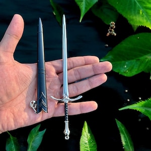 Puede incluir: Una espada plateada en miniatura con empu&ntilde;adura negra y vaina a juego. La espada presenta una guarda detallada y una hoja afilada. La vaina tiene grabados decorativos. La espada mide unos 15 cm de largo.