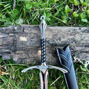 Puede incluir: Una espada decorativa con una hoja y empu&ntilde;adura plateadas, un mango negro y una vaina negra. La espada est&aacute; sobre un trozo de madera, con hierba verde en el fondo.