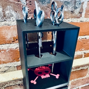 May include: A black, multi-compartment organizer holding several pairs of silver scissors. The bottom compartment features a pink skull and crossbones design. The organizer is set against a brick wall.