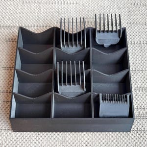 May include: A dark gray organizer with multiple compartments, holding various clipper guards. The organizer has a wavy top edge and is designed to store hair clippers and their attachments. The image is taken from a top-down perspective.