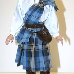 May include: A doll wearing a brown hat, a white shirt, a blue and brown plaid kilt, a brown leather belt with a silver buckle, a brown leather pouch with a silver button, and brown leather boots.