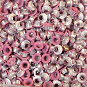May include: A large pile of silver and pink metal eyelets. The eyelets are used for attaching fabric to other materials.