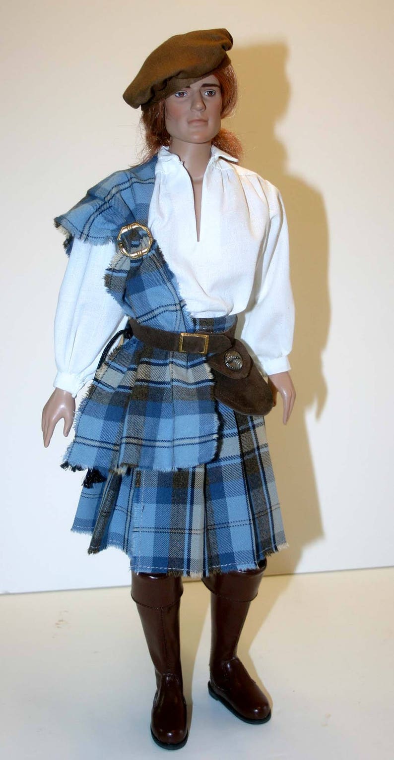 May include: A doll wearing a brown hat, a white shirt, a blue and brown plaid kilt, and brown boots.