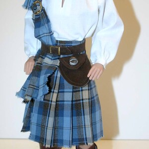 May include: A doll wearing a brown hat, a white shirt, a blue and brown plaid kilt, and brown boots.
