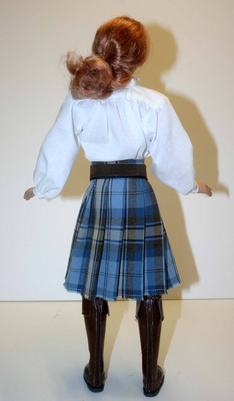 May include: A doll wearing a white blouse, a brown belt, and a blue plaid kilt. The doll is wearing brown boots.