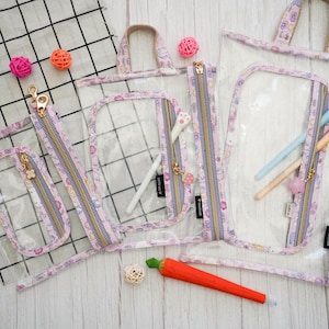 Pode incluir: Quatro bolsas transparentes com zíper de plástico com padrões florais rosa e branco. As bolsas têm zíperes dourados e são de vários tamanhos. A bolsa maior tem uma alça.