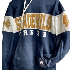 Könnte beinhalten: Marineblauer Kapuzenpullover mit dem Schriftzug "SUNDEVILS CHEER" in goldfarbenem Glitzer auf der Brust. Der Pullover hat ein weißes Band über der Brust und den Ärmeln mit einer goldenen Schleifengrafik. Eine Vordertasche und eine Kapuze mit Kordelzug.