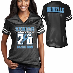 Puede incluir: Camiseta de malla negra con rayas blancas en las mangas. La parte delantera presenta el texto "SENIOR twenty 26 DANCE MOM" en purpurina blanca y azul claro. La parte trasera de la camiseta muestra el nombre "DRINELLE" en purpurina azul claro.