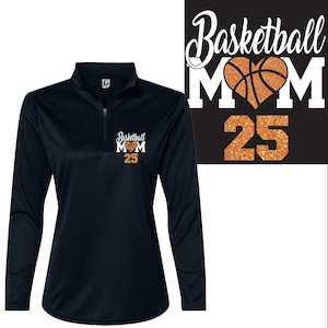 May include: Black long-sleeve athletic shirt with a quarter-zip neckline. The shirt features the text "Basketball MOM 25" in white and orange glitter. A matching design is on a black background.