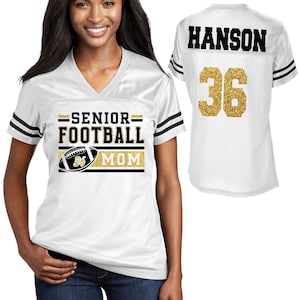 May include: A white V-neck jersey with black stripes on the sleeves. The front features the text SENIOR FOOTBALL MOM and a football graphic with the number 36. The back displays the name HANSON and the number 36 in a gold glitter finish.