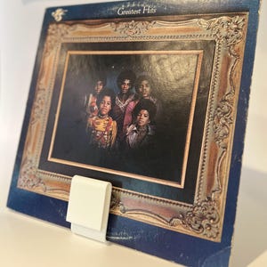 May include: A vintage vinyl record album cover with a gold frame. The cover features a group of five young men in a black and white photo. The album title is "Greatest Hits".