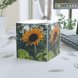 May include: A cube-shaped notepad with a sunflower design. The sunflower is yellow with a dark brown center and green leaves. The background is a blurred image of a garden.