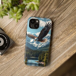 Può includere: Una custodia per telefono con un'aquila calva in volo su un paesaggio montano. Il design include un cielo blu, montagne innevate, un lago e una foresta. L'aquila ha piume marroni e bianche.