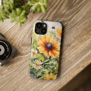 Puede incluir: Una funda para teléfono con un diseño de acuarela de girasoles y abejas. La funda es blanca con la parte posterior transparente y una protuberancia de cámara negra.