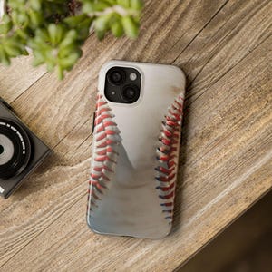 Puede incluir: Una funda blanca para iPhone con diseño de béisbol. La funda presenta una imagen realista de una pelota de béisbol, con costuras rojas y una superficie texturizada. La funda está sobre una superficie de madera.