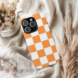 May include: A smartphone case with a vibrant orange and white checkered pattern. The case has a black camera module and a smooth, glossy finish. The design is a classic, eye-catching style.