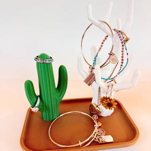 May include: A white ceramic jewelry stand shaped like a deer with antlers, a green cactus ring holder, and a brown tray. The stand has multiple rings and bracelets hanging from it, including a sunflower charm. The tray has a gold bangle bracelet with charms.