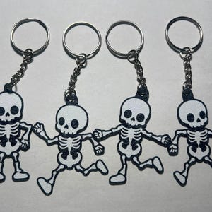 May include: Four keychains featuring white skeleton figures with black accents. Each skeleton has a distinct pose and is attached to a silver-toned metal split ring by a short chain. The skeletons have large, rounded heads and visible rib cages.