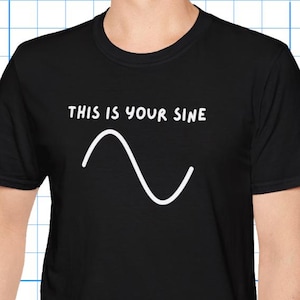 Puede incluir: Camiseta negra con el texto "THIS IS YOUR SINE" sobre un gráfico de onda sinusoidal blanca. La camiseta está hecha de un material suave. La persona lleva vaqueros azules. Adecuada para uso informal.