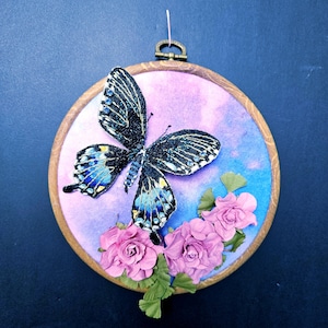 Könnte beinhalten: Gestickter Schmetterling mit schillernden blauen und schwarzen Flügeln, umgeben von rosa Stoffblumen auf einem Aquarellhintergrund in einem hölzernen Stickrahmen.