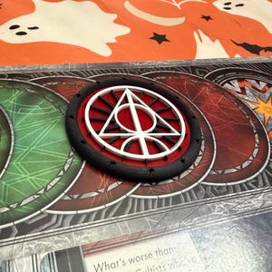 May include: A circular game piece with a white and red symbol, surrounded by a black border with small black shapes. The piece is on a game board with green, red, and gray circular designs. The background is orange with Halloween-themed decorations.