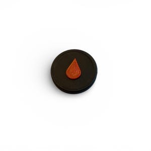 Puede incluir: Un objeto circular negro con una superficie texturizada y una forma de lágrima naranja en relieve en el centro. El objeto está sobre un fondo blanco, creando una imagen de alto contraste.