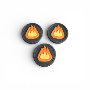 May include: Three circular tokens with a black textured background and a flame design. The flame is orange with a yellow center. The tokens are arranged in a triangular pattern against a white background.