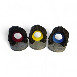 May include: Three game pieces with a cave-like design. Each piece has a black exterior, a gray base, and a colored interior: red, yellow, and blue. The pieces are designed to stand upright.