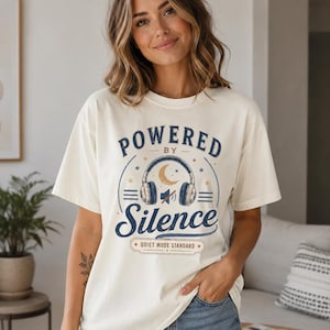 May include: An off-white short-sleeve t-shirt featuring a graphic print with the text "POWERED BY Silence" and "QUIET MODE STANDARD". The design includes an illustration of headphones with a crescent moon and a mute symbol in the center.