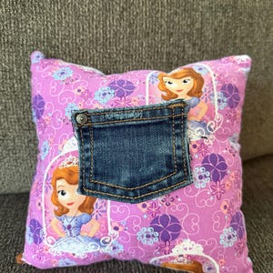 May include: A square throw pillow featuring a purple fabric with a repeating pattern of cartoon princesses in white frames, floral accents, and swirls. A dark blue denim jeans pocket with a metal rivet is sewn onto the center of the pillow.