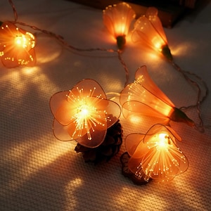 May include: A string of decorative lights featuring translucent orange flower-shaped shades. Each flower contains a bright, warm-toned LED bulb with thin, radiating fiber optic filaments that extend outward from the center, creating a glowing effect.