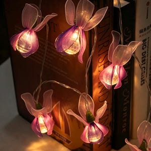 May include: A string of decorative LED lights featuring translucent, orchid-shaped petals in shades of purple and white. Each flower houses a small, warm-glowing bulb, connected by a thin, flexible wire, creating a soft illumination effect.