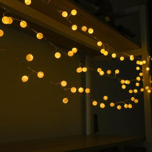 Op de afbeelding: Een decoratieve lichtsnoer met kleine, bolvormige, warmgekleurde lampjes bevestigd aan een dunne, flexibele draad. De lampjes zijn in meerdere gedrapeerde secties gerangschikt en stralen een zacht schijnsel uit in een donkere omgeving.