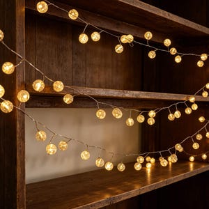 May include: A string of decorative fairy lights featuring numerous small, spherical, translucent bulbs that emit a warm, golden glow. The lights are connected by a thin, flexible wire, creating a continuous line of illumination across the frame.