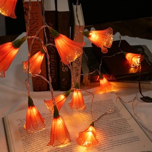 Könnte beinhalten: Eine LED-Lichterkette mit durchscheinenden orangefarbenen blütenförmigen Abdeckungen und grünen Stielen. Die Lichter leuchten in einem warmen Ton und sind über mehrere offene und geschlossene Bücher mit sichtbarem Text auf den Seiten drapiert.