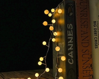 May include: A string of small, glowing warm white spherical LED lights on a thin wire, draped over three books titled Cannes, Sunrise and Sunset, and Big Debt Crises. The lights emit a soft, ambient glow against a dark background.