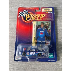 May include: A blue die-cast model of Jeff Gordon's #24 Pepsi Chevrolet Monte Carlo NASCAR race car. The car is in a clear plastic package with a red, blue, and white backing featuring the "Winner's Circle" logo and a photo of Jeff Gordon. The packaging includes the text "Lifetime Series".