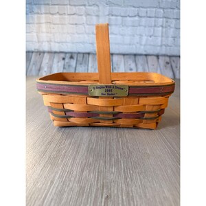 May include: A rectangular woven basket with a wooden handle. The basket is made of light brown and dark brown woven strips. A metal plaque on the front reads "It Begins With A Dream" and "1995 Bee Basket".