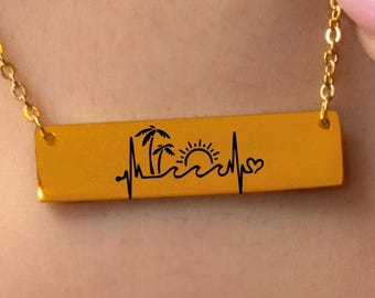May include: Gold-colored necklace with a rectangular pendant. The pendant features a black graphic of palm trees, a sun, waves, and a heartbeat line ending in a heart shape. The necklace is on a gold chain.