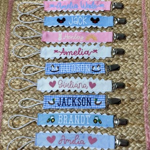May include: A collection of baby pacifier clips in various colors, each with a name embroidered on a decorative ribbon. The clips feature names like Amelia, Jack, and Hudson, with decorative elements such as hearts, ducks, and tractors.