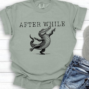 May include: Sage green t-shirt featuring the text "AFTER WHILE" above a black illustration of a dancing alligator. The alligator is depicted with its arms raised and its tail curved. The shirt is made of a soft material.