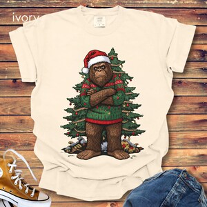 May include: An ivory t-shirt featuring a graphic of a brown Bigfoot wearing a red Santa hat and a green patterned sweater with arms crossed. Behind the creature is a decorated Christmas tree with red ornaments and gold garland, with gifts at the base.