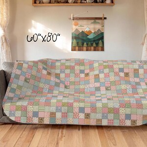 Puede incluir: Un edredón de retazos, de aproximadamente 152 cm x 203 cm, cubriendo un sofá. El edredón presenta una variedad de cuadrados de colores pastel con patrones florales y a cuadros. Un adorno de pared de madera con una escena de montaña es visible en el fondo.