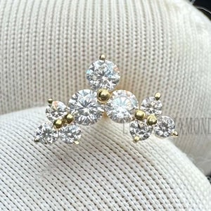 Könnte beinhalten: Ein Cluster aus funkelnden Diamanten, gefasst in einer goldenen Basis. Das Design zeigt mehrere rund geschliffene Diamanten, die in einem floralen oder sternförmigen Muster angeordnet sind und einen schillernden Effekt erzeugen. Der Schmuck wird auf weißem Stoff präsentiert.