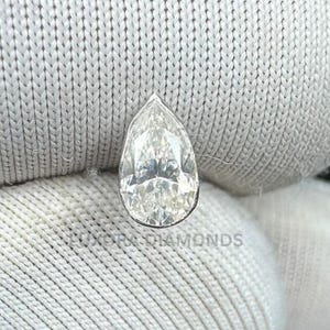 May include: A pear-shaped diamond with a brilliant cut, set against a white fabric background. The diamond is clear and reflects light, showcasing its facets. The image is well-lit, highlighting the diamond's clarity and shape. The text "Luxury Diamonds" is visible.