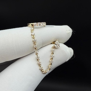 Puede incluir: Un puño de oreja de oro con una cadena y un diamante en forma de pera colgante. El puño de oreja tiene una fila de pequeños diamantes. La cadena está adornada con pequeños diamantes. El texto "LUXORADIAMONDS" es visible.