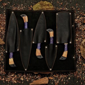 Op de afbeelding: Een set van vijf messen met houten handvatten en paarse accenten, elk in een zwarte leren schede. De messen zijn gerangschikt in een zwarte leren etui met een donkere houten achtergrond, omringd door bladeren en houtkrullen.
