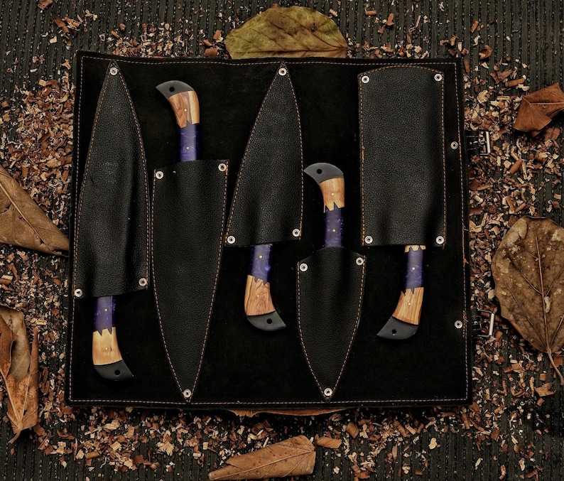 Op de afbeelding: Een set van vijf messen met houten handvatten en paarse accenten, elk in een zwarte leren schede. De messen zijn gerangschikt in een zwarte leren etui met een donkere houten achtergrond, omringd door bladeren en houtkrullen.