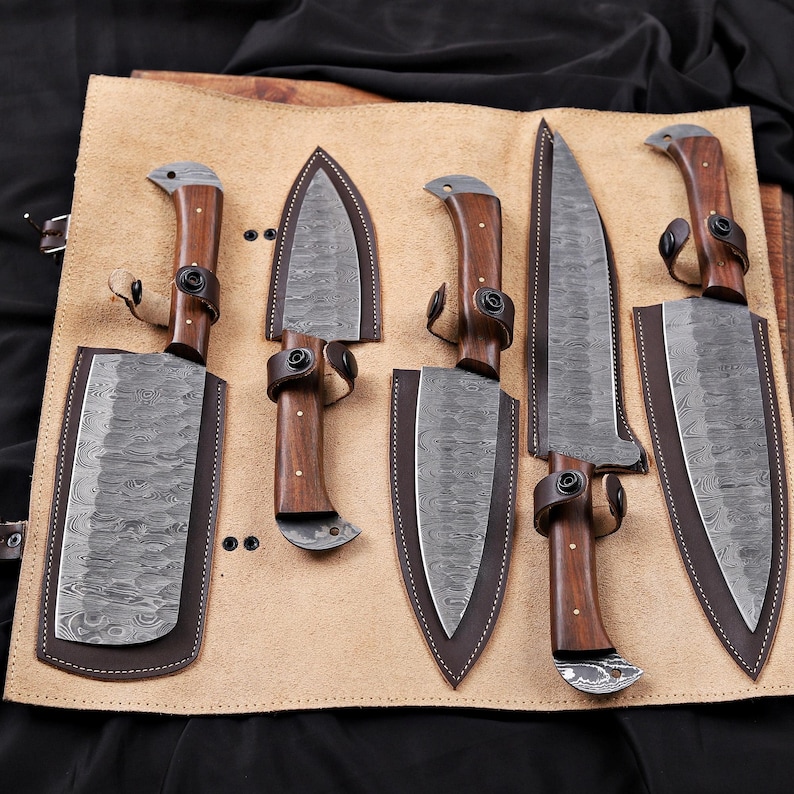 Op de afbeelding: Een set van zes messen met houten handvatten en patronen op de bladen, gepresenteerd op een bruinleren rol. Elk mes heeft een leren schede en een donkerbruin handvat. De bladen hebben een uniek, getextureerd patroon.